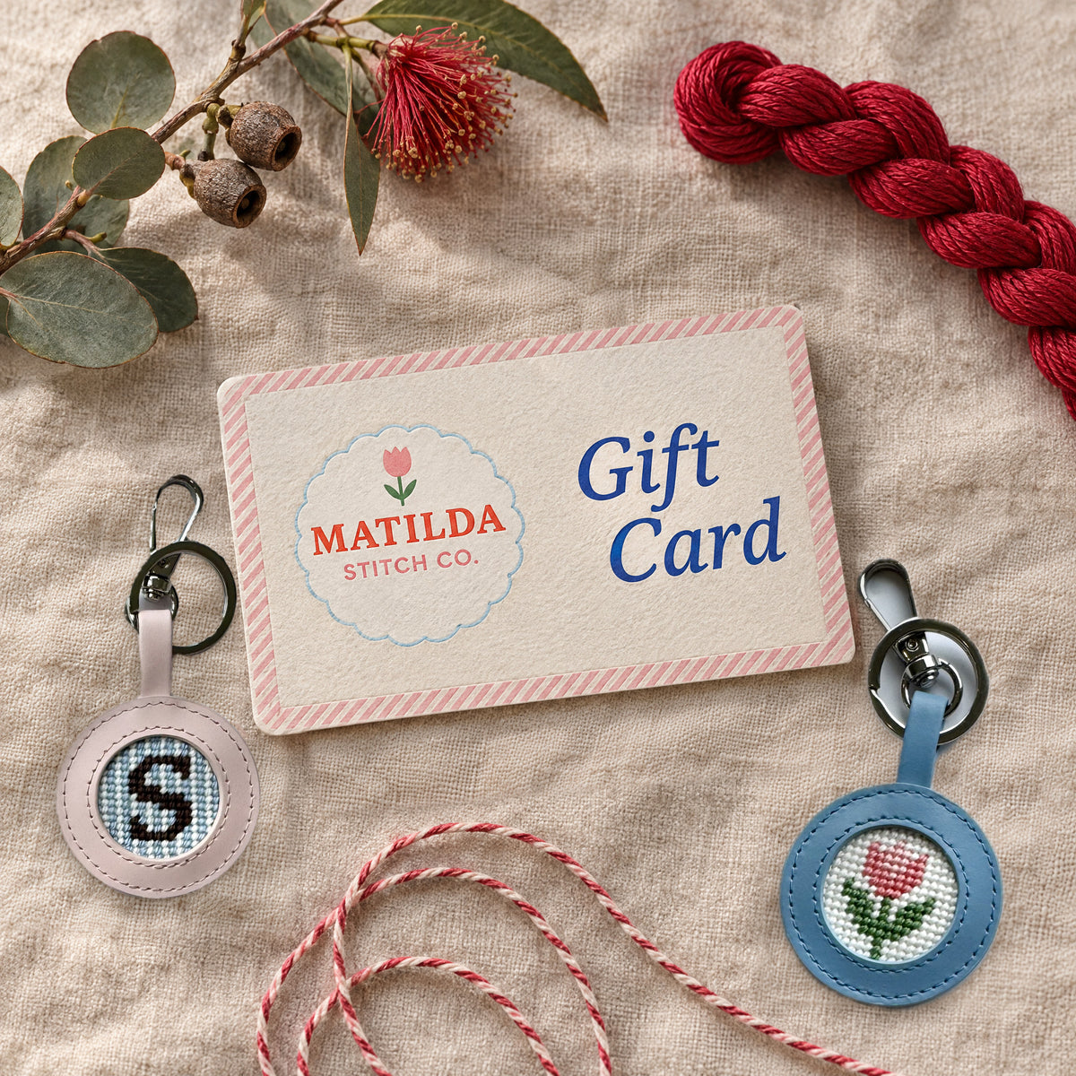 A letterpress Matilda Stitch Co gift card on a linen table with a stitched S initial needlepoint keyring, a stitched tulip needlepoint keyring, a skein of red thread, eucalyptus with gumnuts, red flowering gum and red and white twine.